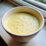 A creamy, yellow Summer Corn And Zucchini Soup served hot in a white bowl near a window.