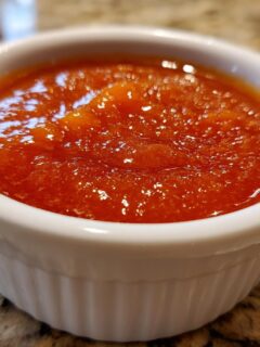 A close-up of vibrant orange-red Sweet And Sour Sauce in a small white ramekin on a granite counter.