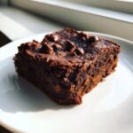 A close-up of a rich, dark chocolate slice of Sweet Potato Brownies topped with chocolate chips on a white plate.