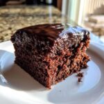 Close-up of a rich, dark slice of Texas Sheet Cake topped with glossy chocolate frosting on a white plate.