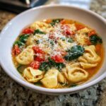 A close-up of a white bowl filled with steaming Tortellini Soup, featuring cheese tortellini, spinach, tomatoes, and grated Parmesan.