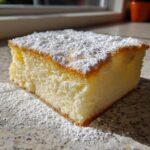 A close-up, sunlit slice of light, airy Tres Leches Cake dusted heavily with powdered sugar.