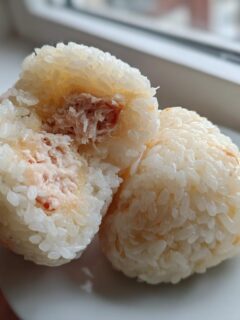 A hand holding two Tuna Mayo Rice Balls, one broken open to show the creamy tuna filling inside the sticky rice.