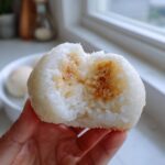 A hand holding a Tuna Mayo Rice Balls ball broken open to show the savory, brownish-orange filling inside the white rice.