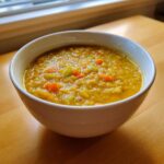 A close-up of a white bowl filled with thick, yellow Lentil Soup featuring visible chunks of carrots and celery.