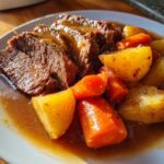 Close-up of tender sliced Crockpot Roast covered in rich gravy, served with chunks of carrots and potatoes.