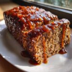 Close-up of a thick slice of savory meatloaf covered in a thick, shiny Bourbon Glaze Meatloaf sauce dripping onto a white plate.
