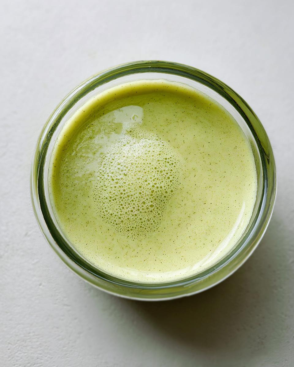 Overhead view of a bright green, slightly frothy liquid, possibly a wasabi-infused drink, in a clear glass.