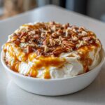 A white bowl filled with creamy Caramel Apple Dip, drizzled heavily with caramel sauce and topped with chopped pecans.