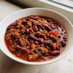 A close-up of a white bowl filled with rich, meaty Chili Recipe Crockpot featuring kidney beans and black beans in a savory red sauce.