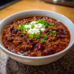Close-up of a white bowl filled with rich, thick Crockpot Chili topped with sour cream and fresh green onions.
