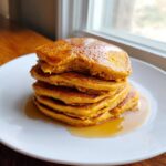 A tall stack of fluffy Pumpkin Pancakes drizzled with syrup on a white plate.