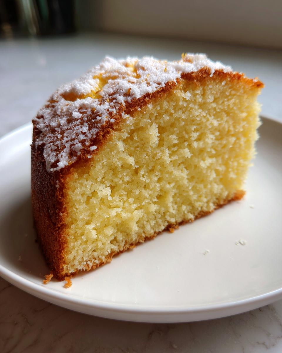 Close-up of a moist slice of yellow vegan cake topped with powdered sugar on a white plate.