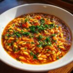 Close-up of a white bowl filled with rich, tomato-based Greek Manestra pasta, topped generously with fresh chopped parsley.