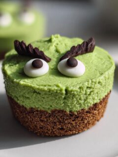 A single Grinch Mini Cheesecake featuring a green top layer, brown crust, and decorative white and brown eyes with chocolate eyebrows.