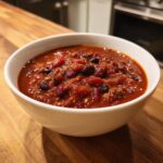 A close-up of a white bowl filled with rich, thick Healthy Vegetarian Three Bean Chili on a wooden counter.