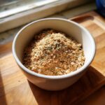 A close-up of homemade Onion Soup Mix, a coarse brown powder with dried herbs, sitting in a small white bowl.