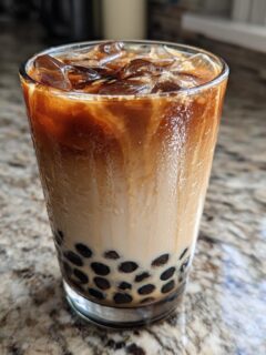 A tall glass of iced coffee bubble tea with tapioca pearls settling at the bottom, showing layers of milk and dark coffee.