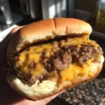 Close-up of a person holding a juicy, cheesy Stuffed Burgers patty melting out from a soft bun.
