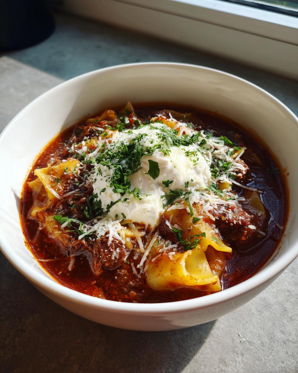 A close-up of a white bowl filled with rich Lasagna Soup Crockpot, topped with a dollop of ricotta, shredded Parmesan, and fresh parsley.