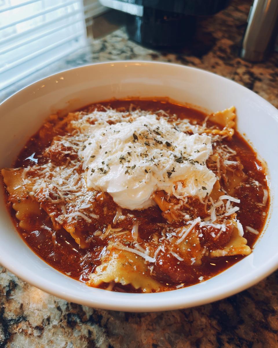 A white bowl filled with Lasagna Soup Crockpot featuring ravioli, topped with ricotta cheese, parmesan, and herbs.