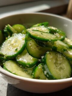 Close-up of thinly sliced, marinated cucumbers seasoned with herbs and salt in a white bowl, illustrating fresh Cucumber Recipes.