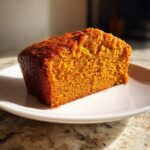 A thick, moist slice of bright orange Pumpkin Cake resting on a white plate, highlighted by sunlight.