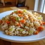 A close-up of a serving of flavorful brown rice recipes mixed with diced carrots and celery on a white plate.