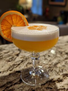 A vibrant orange Sour Cocktails recipe, topped with white foam and garnished with an orange slice.