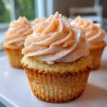 A close-up photo of a freshly baked Peach Bellini Cupcakes featuring a golden cake base and light peach-colored buttercream frosting.