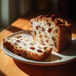 A slice of homemade fruit bread, rich with raisins, sits next to the remaining loaf on a plate, highlighting the texture of these fruit bread recipes.