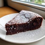 A decadent slice of dark chocolate Flourless Cakes dusted generously with powdered sugar on a white plate.