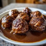 Close-up of several glossy Salisbury Steak Meatballs submerged in a rich brown gravy on a white plate.