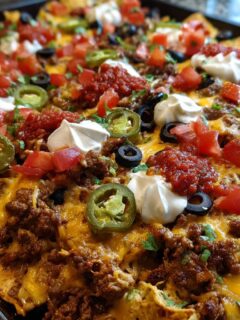 Close-up of fully loaded Sheet Pan Nachos covered in ground beef, melted cheese, sour cream, tomatoes, olives, and jalape&ntilde;os.
