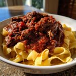 A close-up of tender shredded beef Short Rib Ragu generously spooned over wide, fresh pappardelle pasta in a white bowl.