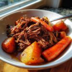 A white bowl filled with tender, shredded Crockpot Pot Roast, large chunks of carrots, and potatoes in rich gravy.
