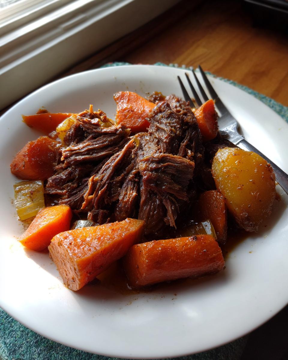 A serving of tender, shredded Crockpot Pot Roast served with chunks of cooked carrots and potatoes on a white plate.