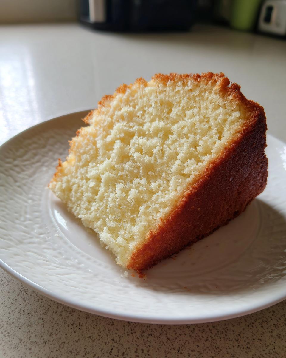 A close-up of a moist, light yellow slice of vegan cake with a golden-brown crust on a white plate.