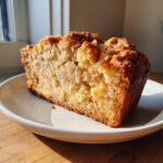 A close-up slice of moist Apple Bread featuring visible apple chunks and a golden brown crumb topping, served on a white plate.