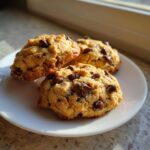 Three soft Pumpkin Chocolate Chip Cookies piled slightly on a white plate, illuminated by natural light.
