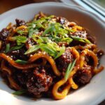 A close-up, appetizing photo of Sticky Beef Noodles coated in dark sauce, topped with green onions and sesame seeds.