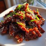 A close-up of crispy, glazed Sticky Orange Beef pieces garnished with sesame seeds and sliced green onions.