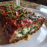 A thick slice of Stuffed Italian Meatloaf showing a melted mozzarella and herb filling, topped with tomato glaze and parsley.