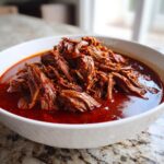 A white bowl filled with rich, deep red broth and a generous mound of shredded, tender meat from Traditional Mexican Birria.
