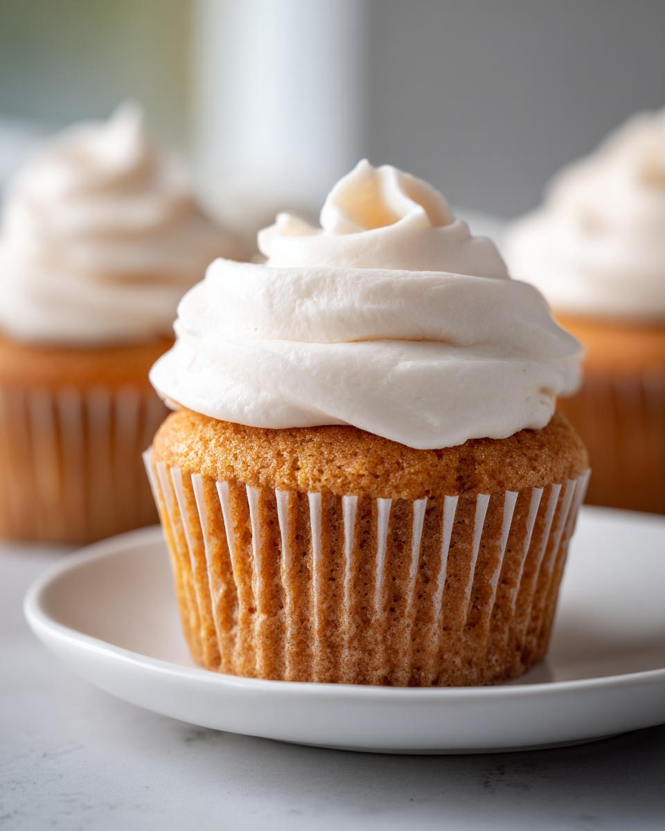 A close-up of one perfect vanilla Sugar Free Cupcakes topped with a swirl of white, creamy frosting, sitting on a small white plate.