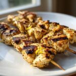 Close-up of grilled Zesty Parmesan Lemon Chicken Skewers showing char marks and sprinkled lemon zest on a white plate.