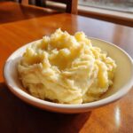A close-up of a bowl filled with fluffy, creamy Best Mashed Potatoes sitting on a sunlit wooden table.