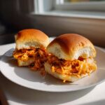 Two freshly made Buffalo Chicken Sliders filled with shredded, saucy chicken and melted cheese, served on a white plate.