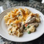 A serving of creamy Chicken Pot Pie Pasta topped with melted cheddar cheese, peas, and carrots on a white plate.