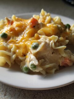 A close-up of a serving of creamy Chicken Pot Pie Pasta topped with melted cheddar cheese, peas, and carrots.